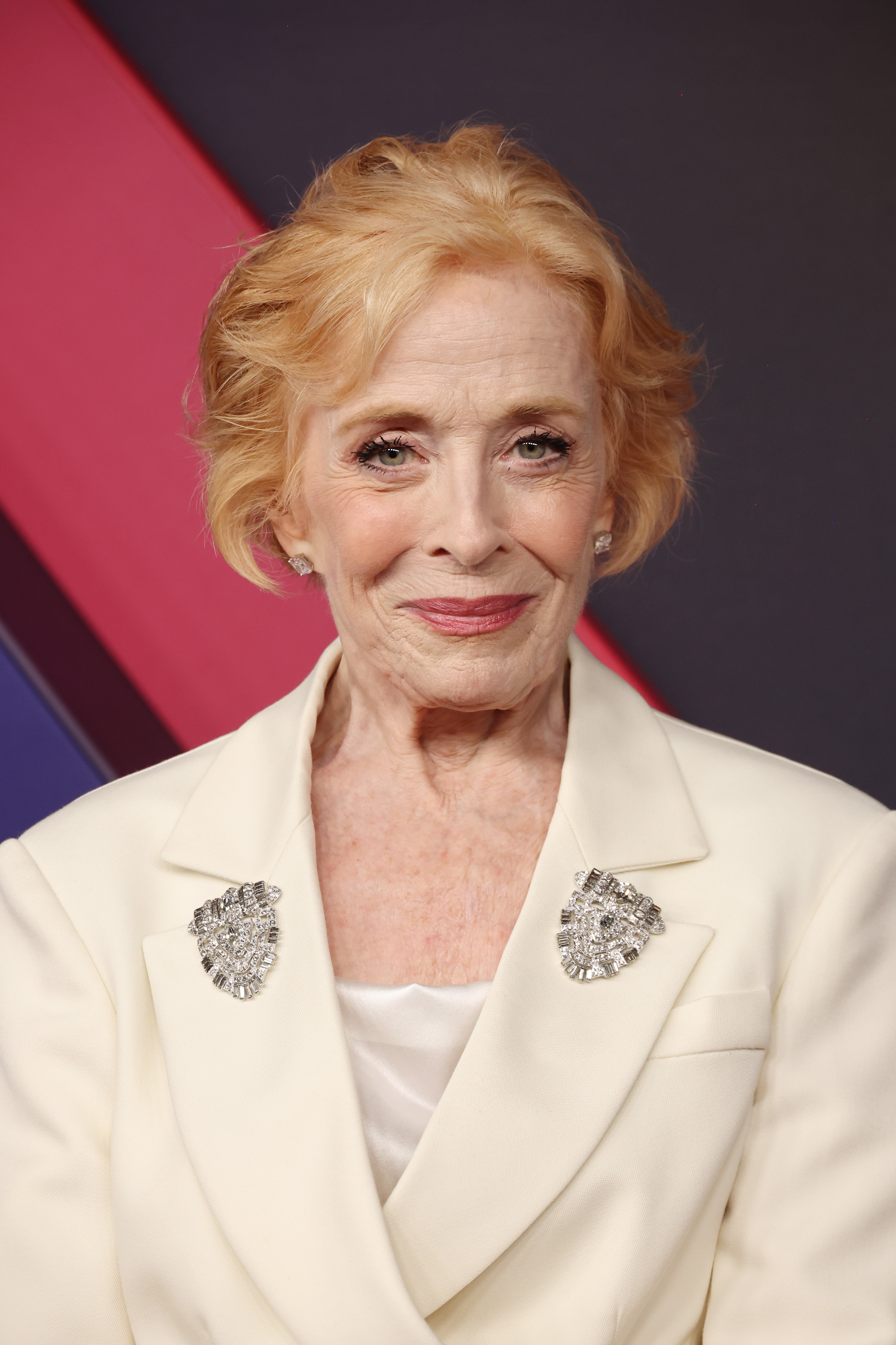 Holland Taylor poses on the red carpet, wearing a white blazer adorned with jeweled brooches
