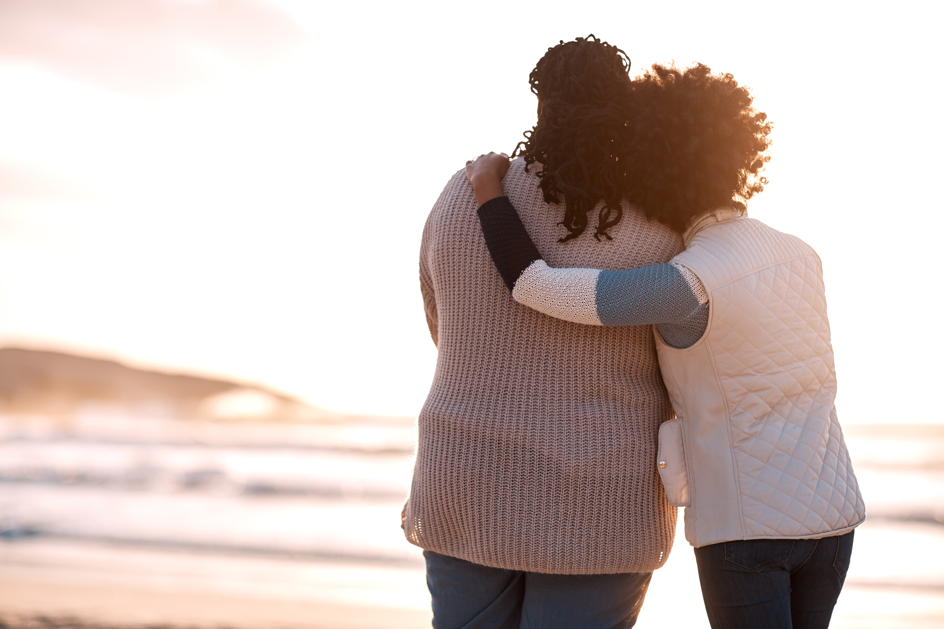 Two people in knitwear stand arm-in-arm, facing the ocean at sunset. Both are in warm, casual vests