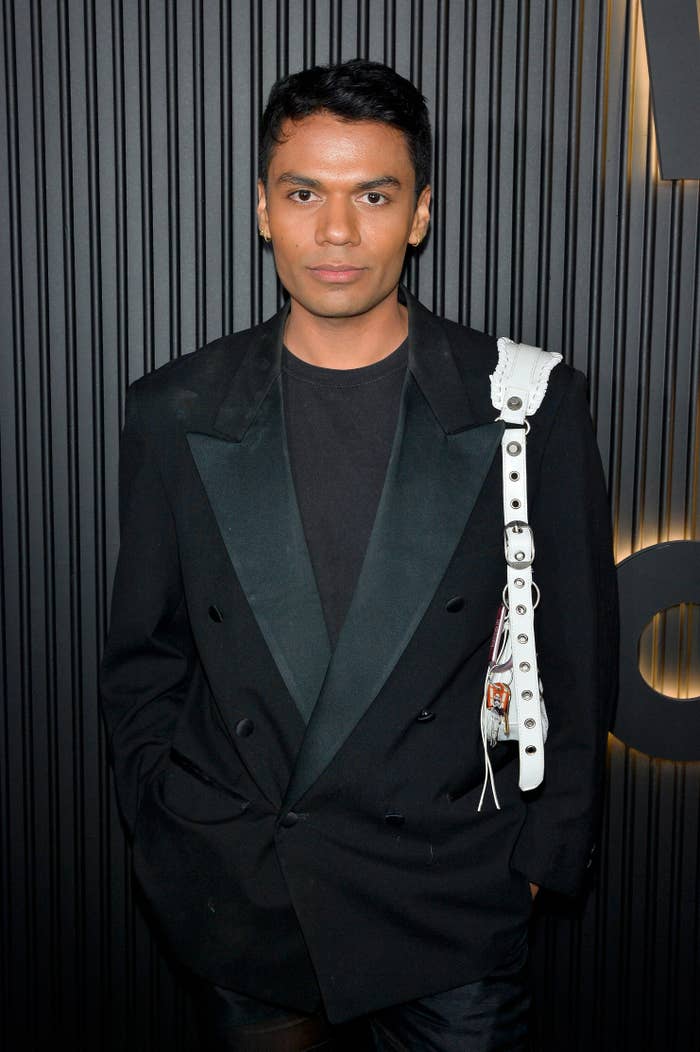 Person wearing a black blazer and black shirt with a white strap across the shoulder stands in front of a patterned wall