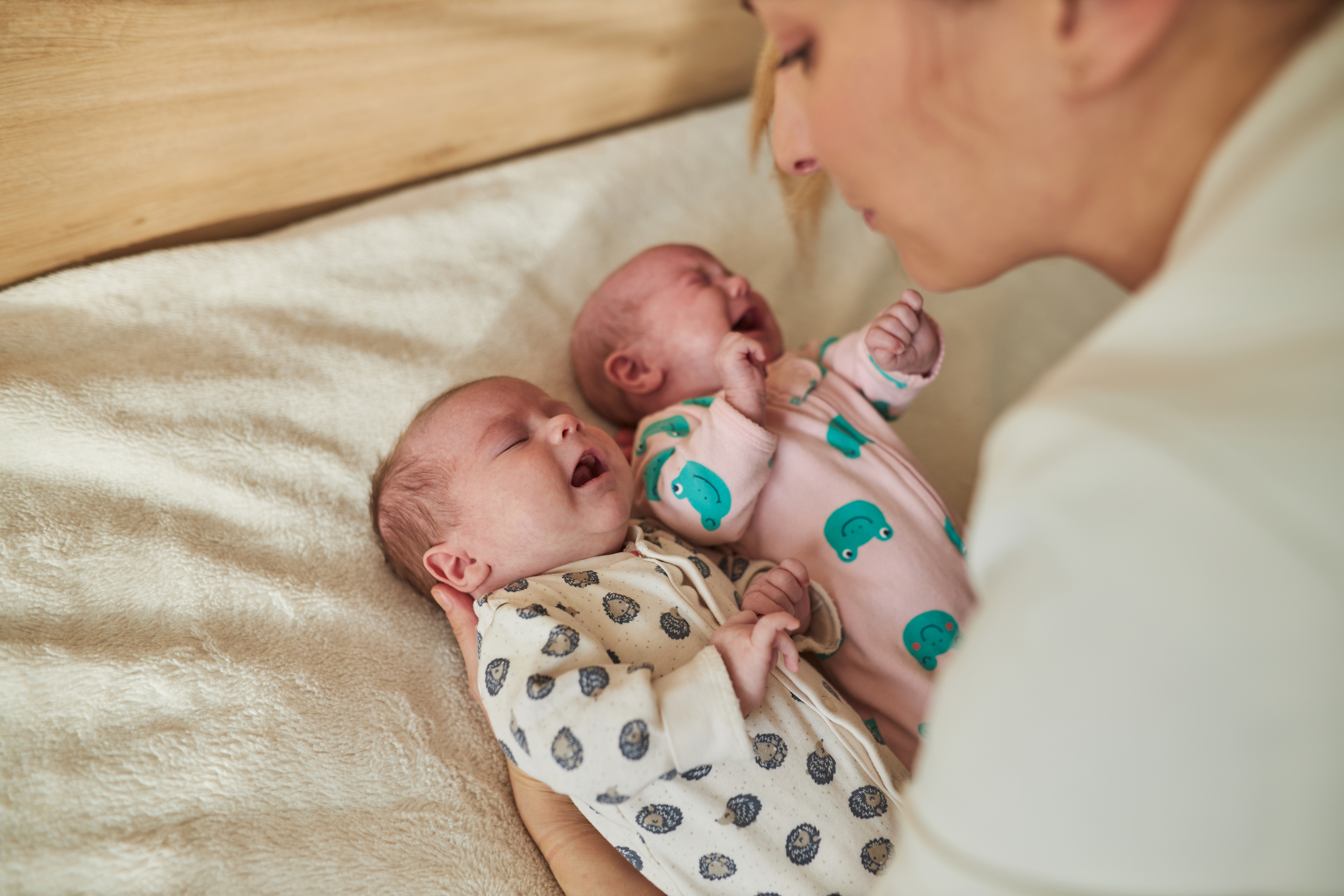 An adult comforts two newborn babies lying on a bed, one dressed in a polka dot outfit and the other in an outfit with elephant prints