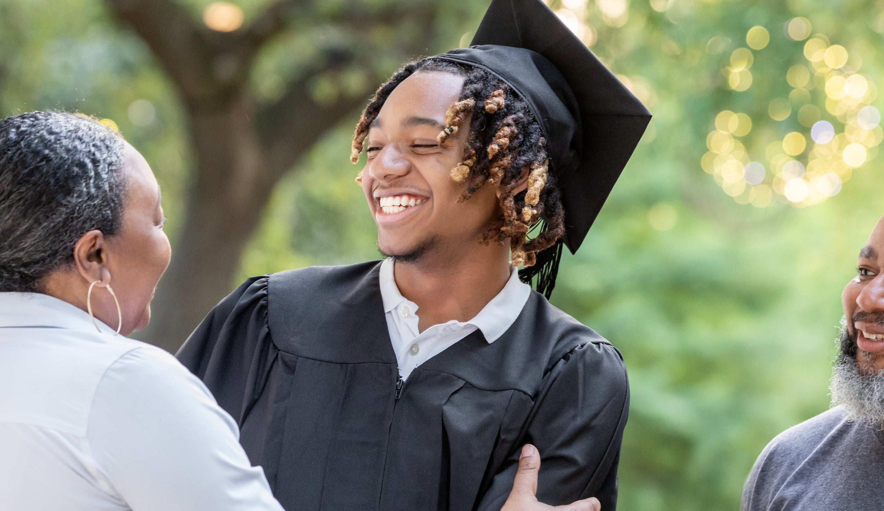 A young male graduate in a cap and gown smiles while being hugged by a woman as a man stands beside them, all appearing happy outdoors. Names not provided