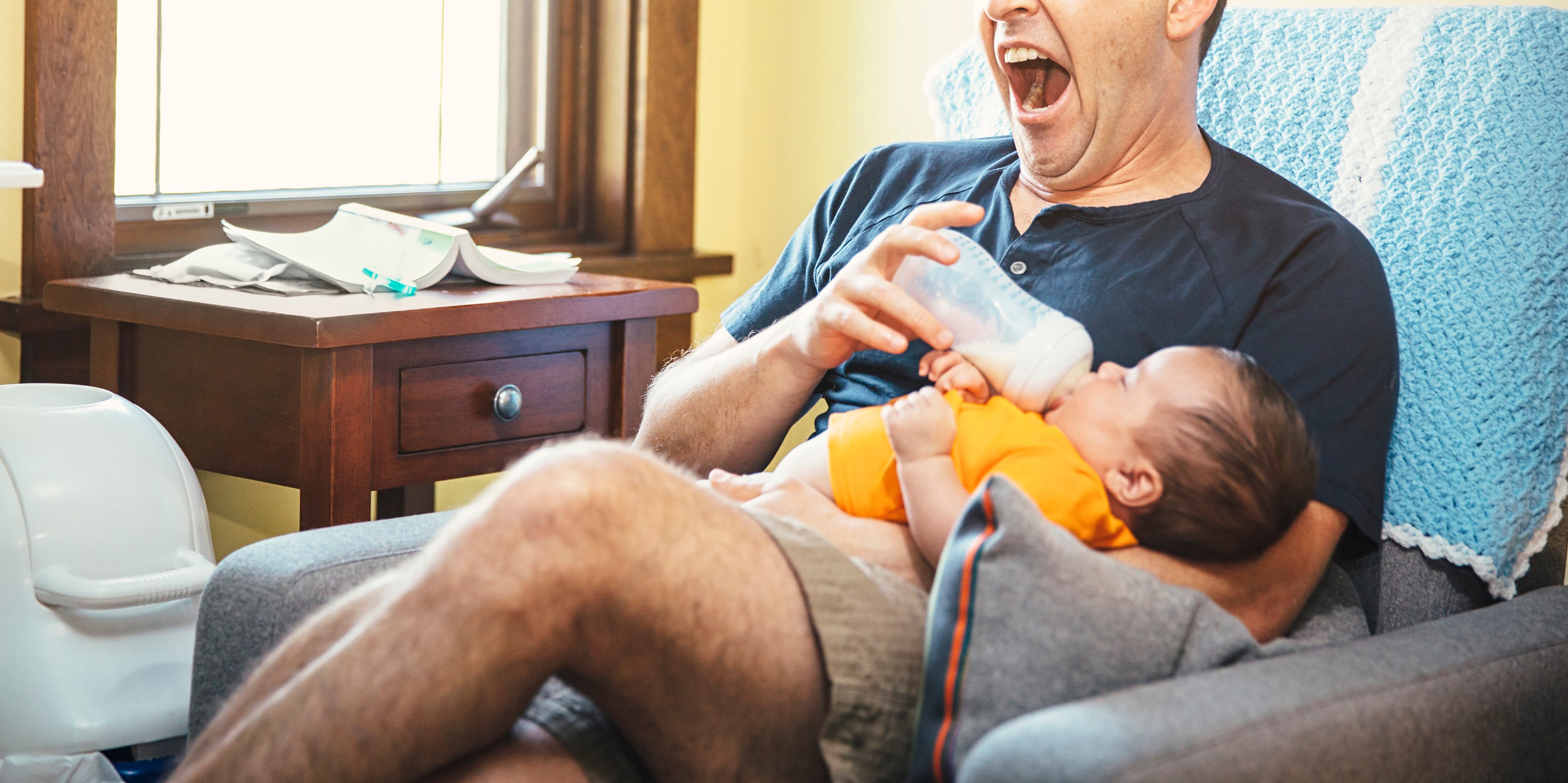 A man yawns while bottle-feeding an infant, sitting in a cozy room with natural light coming from a nearby window