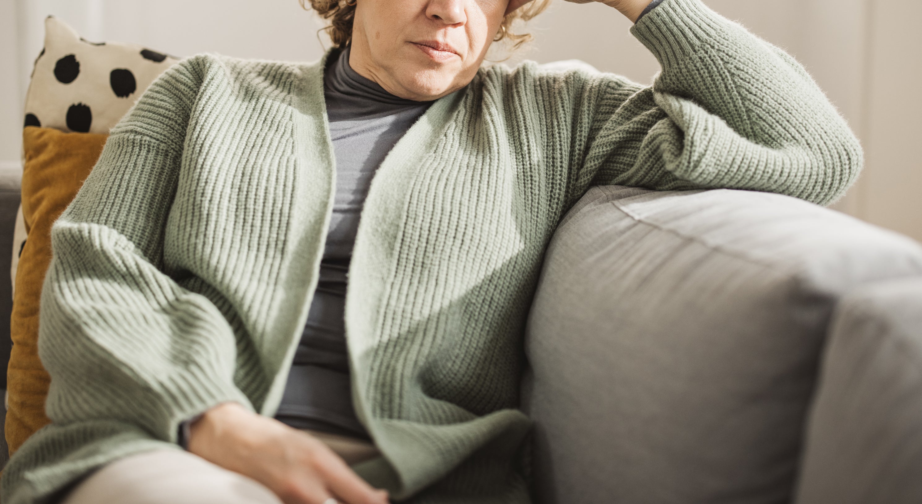 A woman with curly hair sits on a couch, looking thoughtful and resting her head on her hand, wearing a knit cardigan and a turtleneck