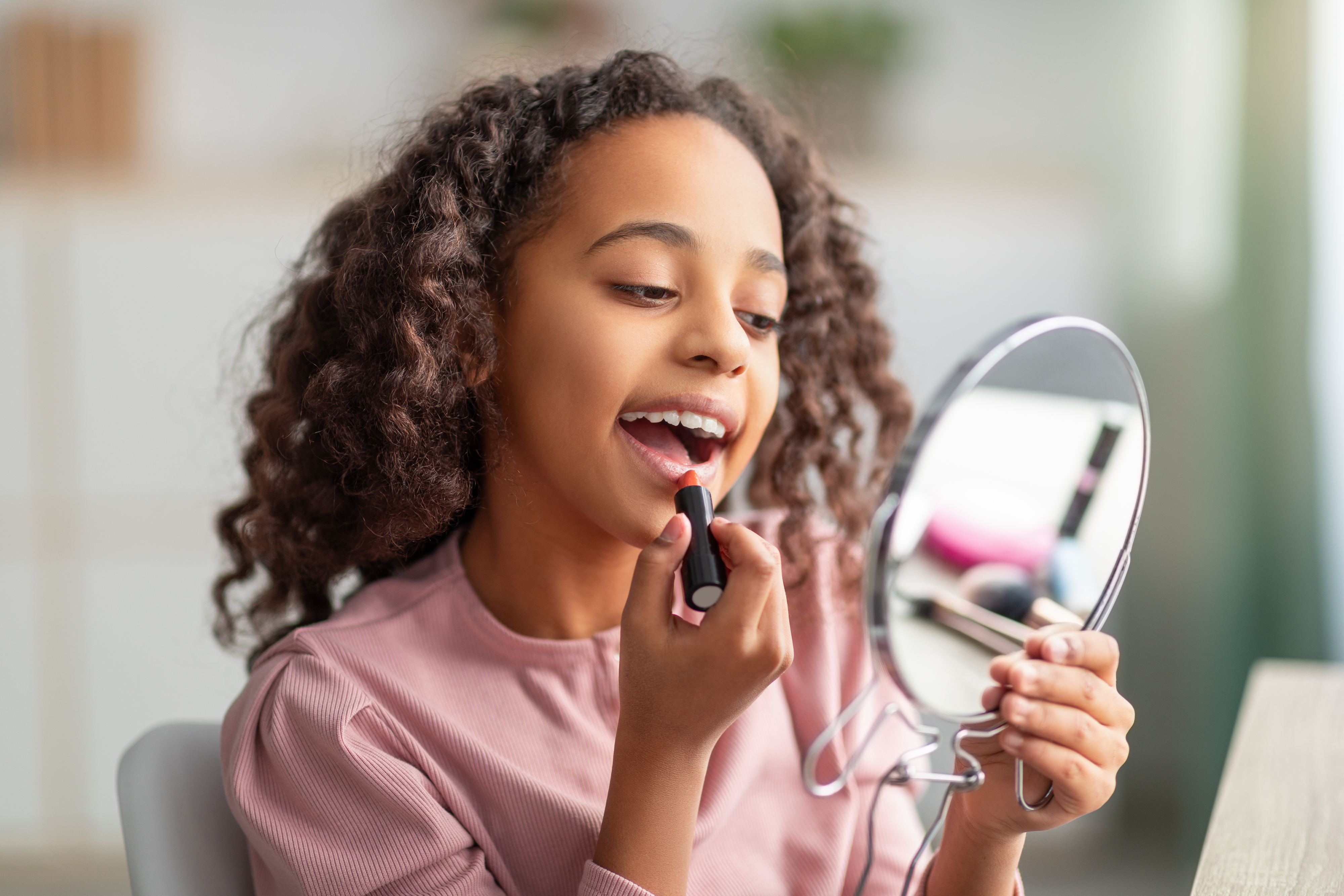 A young girl is applying lipstick while looking into a handheld mirror. She has curly hair and is smiling