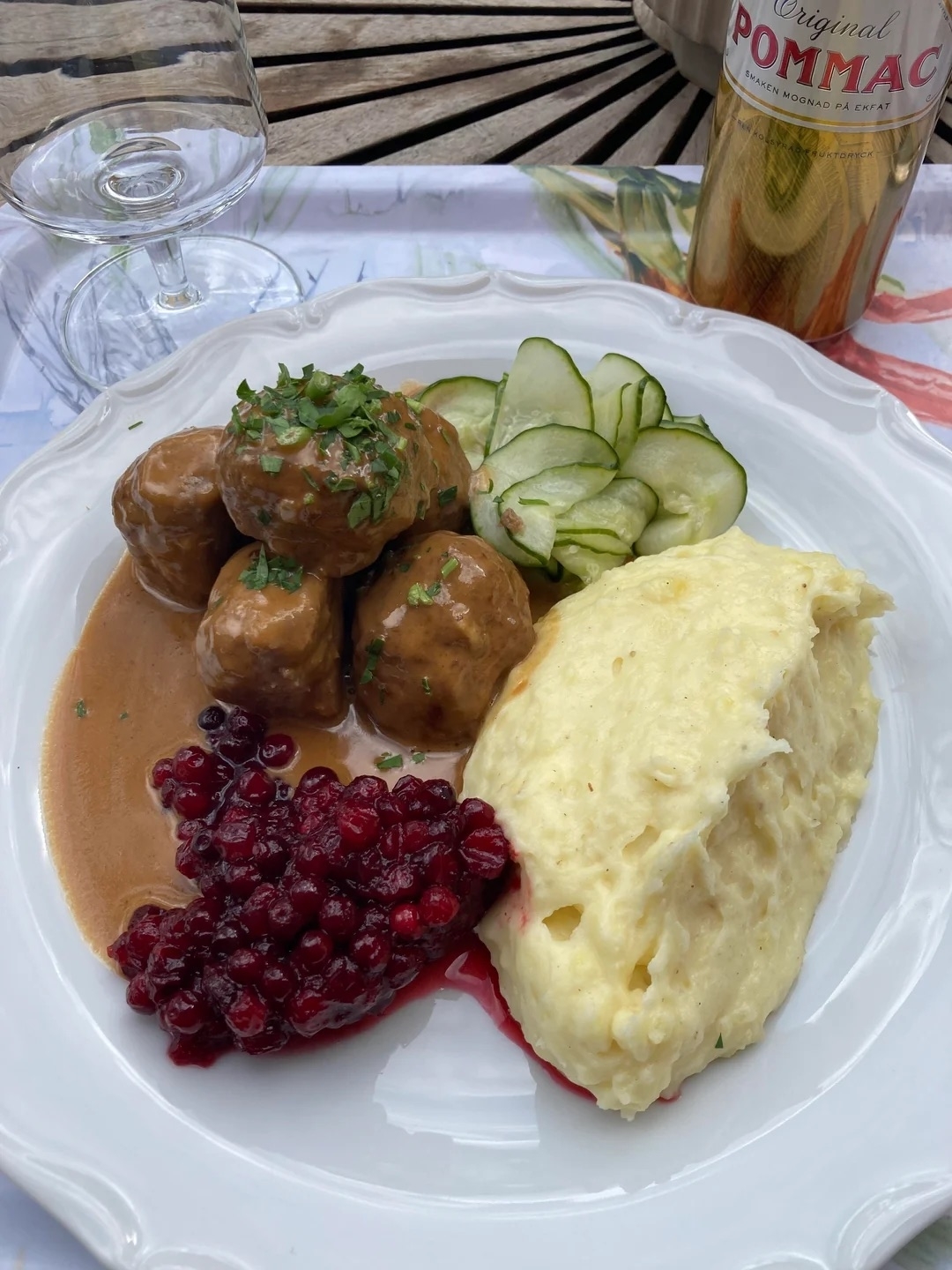Plate with Swedish meatballs in gravy, mashed potatoes, lingonberries, cucumbers, an empty glass, and a bottle of Pommac soda