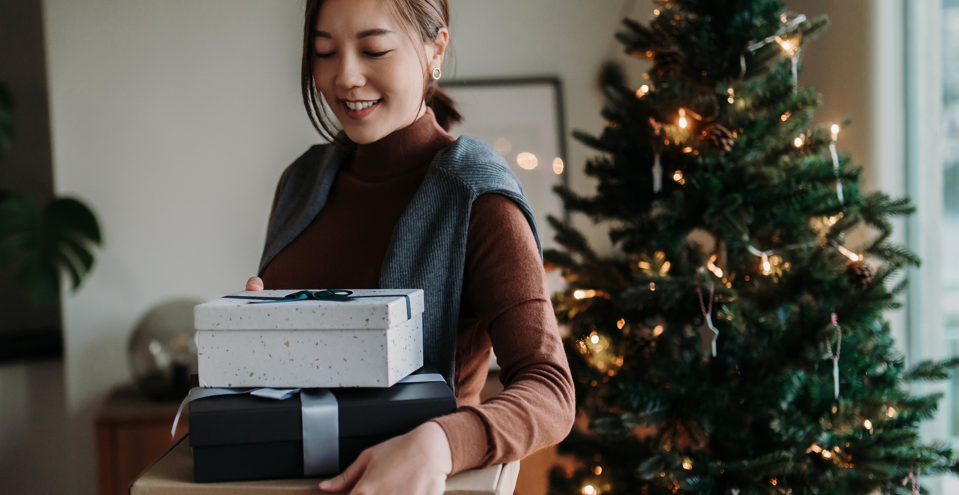 A woman smiles while holding several gift boxes near a decorated Christmas tree with lights. Unknown woman