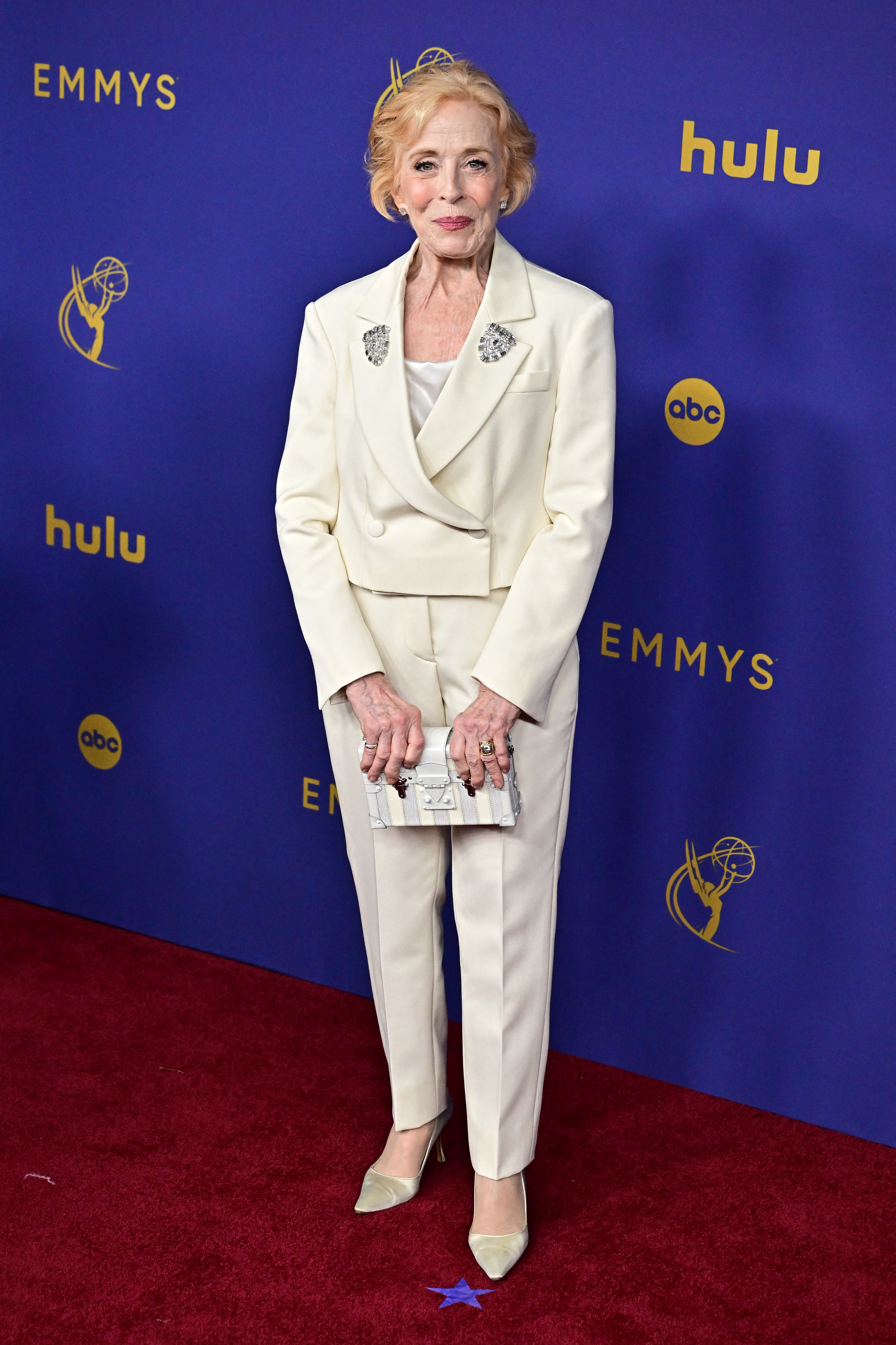 Holland Taylor on the red carpet in a tailored white pantsuit at the Emmys, holding a small clutch