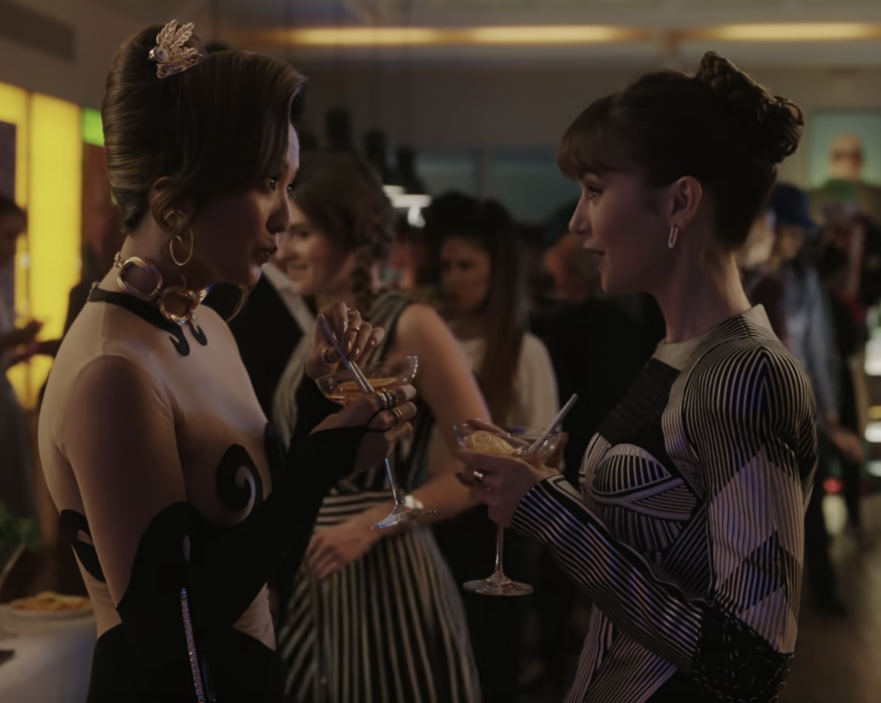 Ashley Park and Lily Collins in stylish cocktail attire, holding martini glasses and engaging in conversation at a lively social event