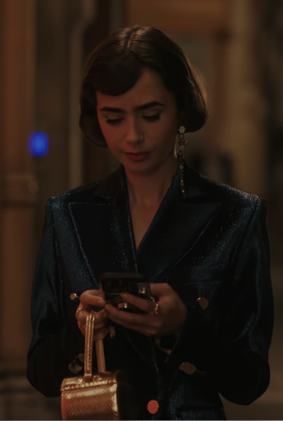 Lily Collins looks at her phone while holding a small purse, wearing a shiny, structured blazer with prominent earrings