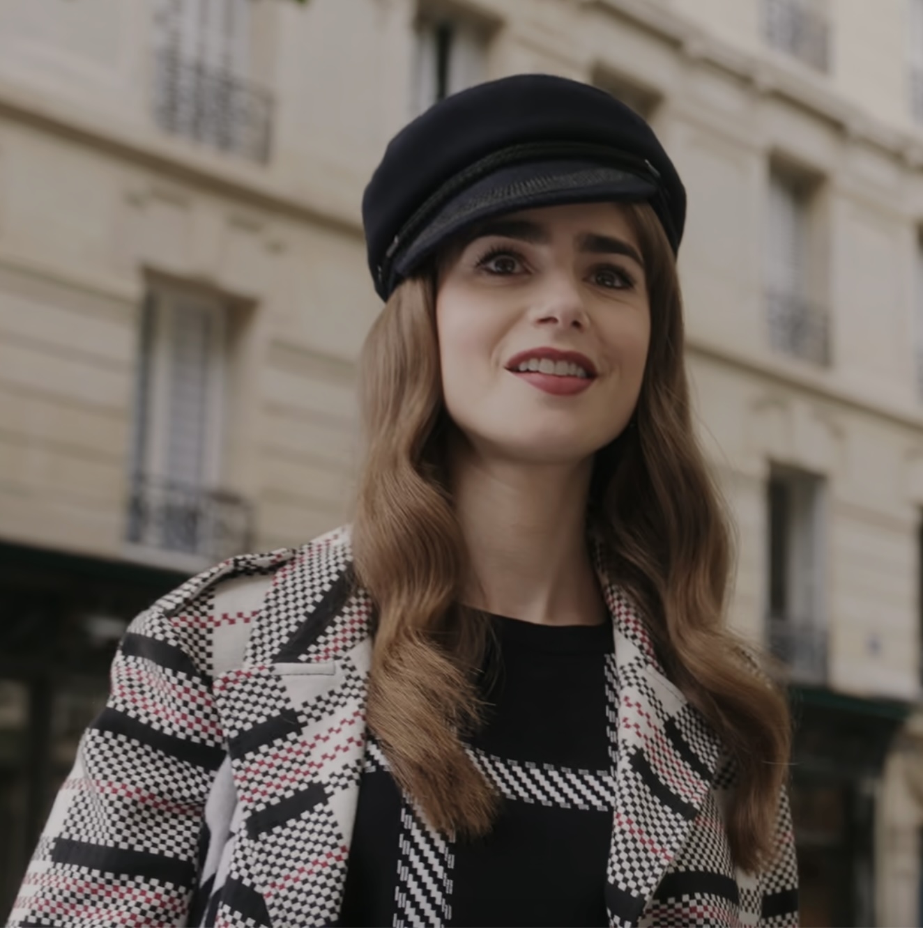 Lily Collins outdoors, smiling, wearing a patterned coat and a stylish hat in a cityscape background