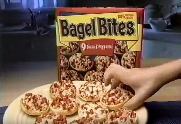 Box of Bagel Bites with a plate of prepared Bagel Bites; a hand reaches for one. The box shows "9 Cheese &amp;amp; Pepperoni"