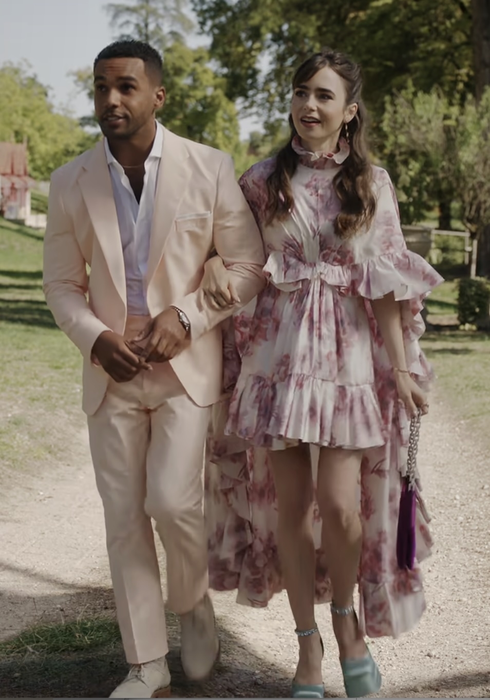 Lucien Laviscount in a light suit and Lily Collins in a flowing floral dress, walking arm-in-arm outdoors
