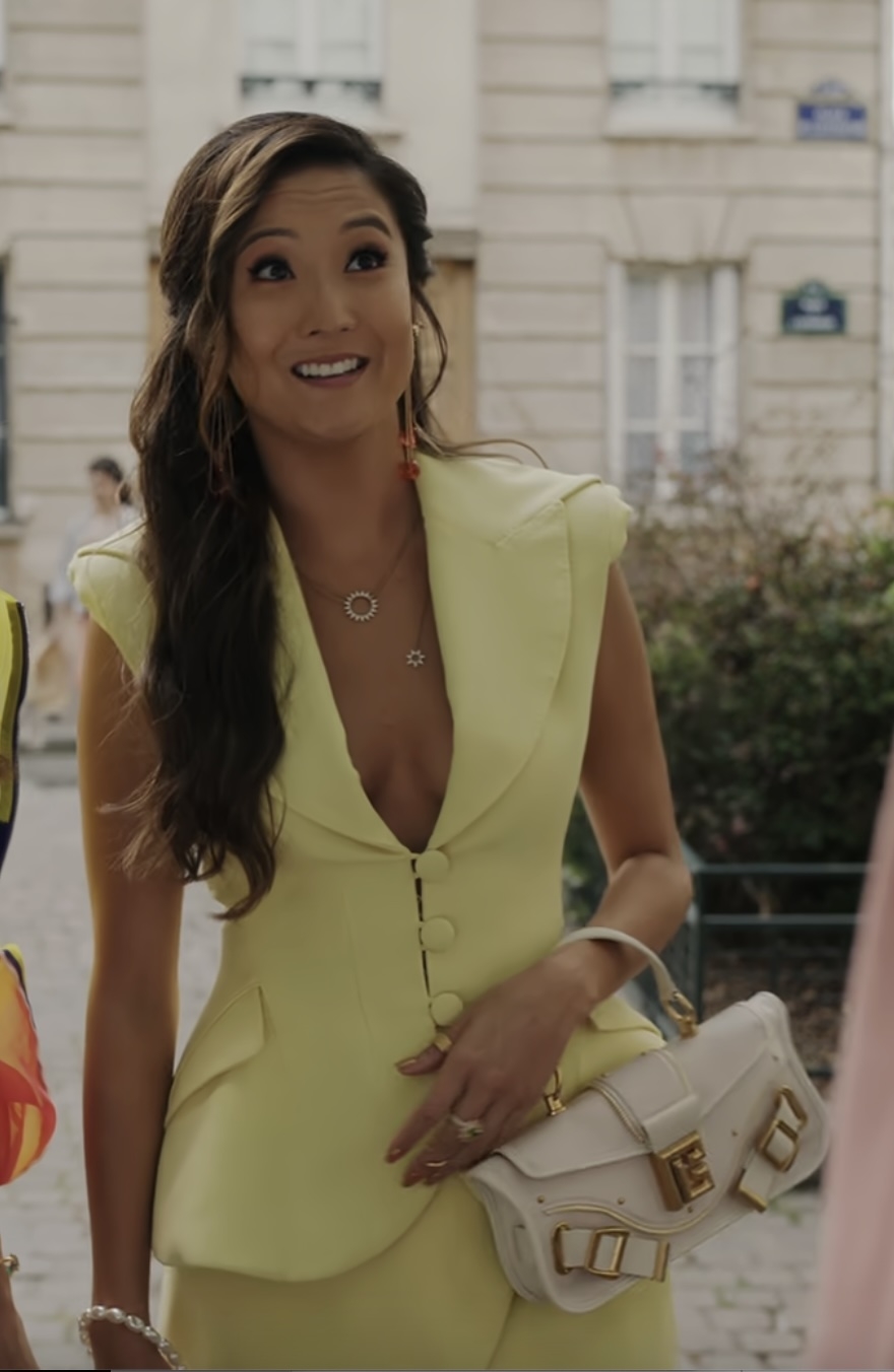 Ashley Park on a street, wearing a fashionable V-neck yellow suit with a matching vest and pants, holding a white handbag