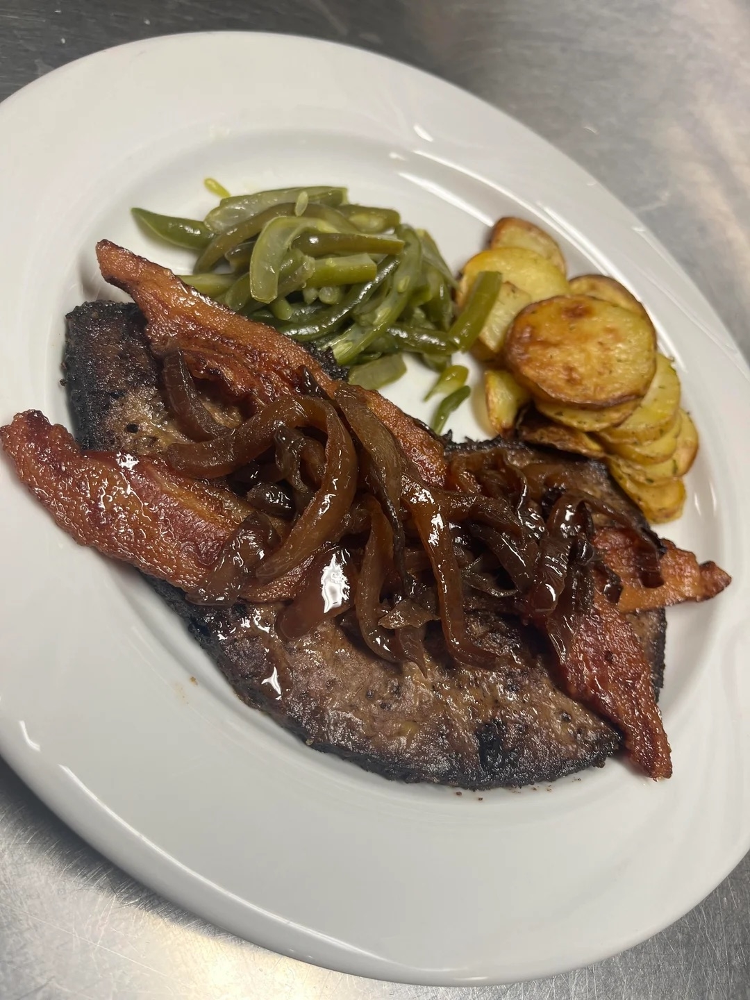 A plate with cooked liver topped with caramelized onions and bacon, with sides of sautéed green beans and roasted potato slices