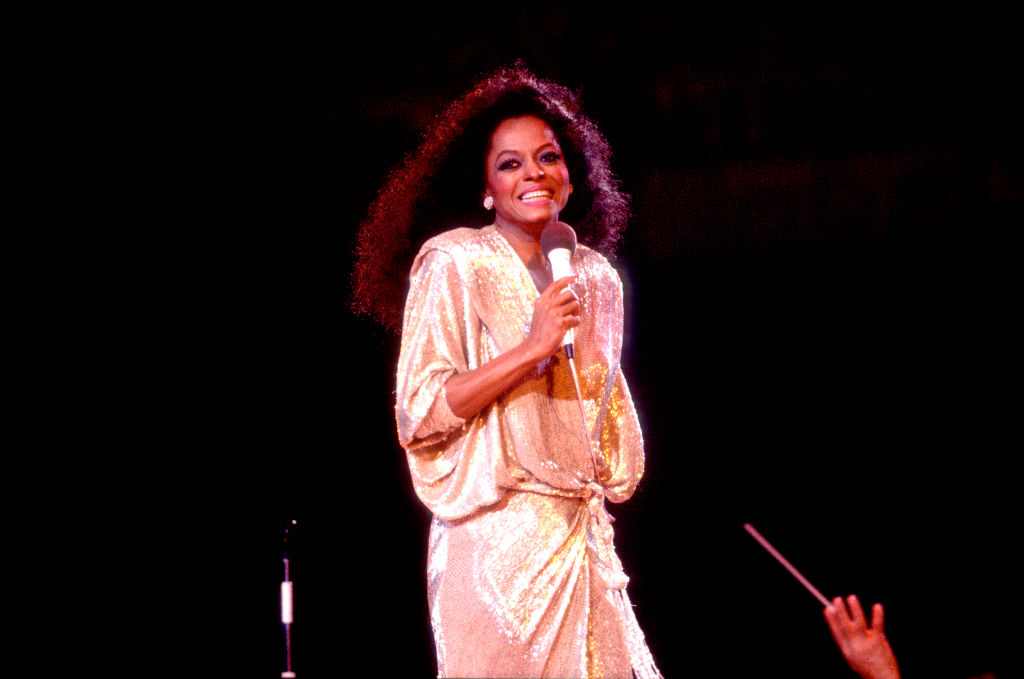 Diana Ross wearing a shimmering gown, performs while holding a microphone, smiling on stage