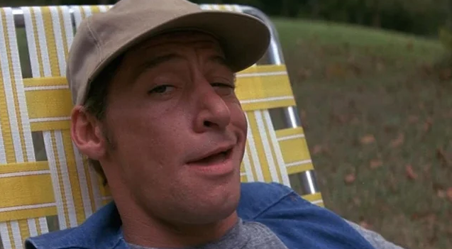 Jim Varney reclines on a striped outdoor lounge chair, smirking with a sideways glance and wearing a cap, outdoor setting in the background