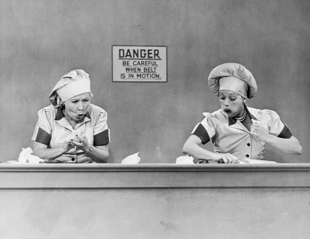 Lucille Ball and Vivian Vance as factory workers, wearing uniforms and headscarves, struggle to keep up with a fast-moving conveyor belt in a comedy scene