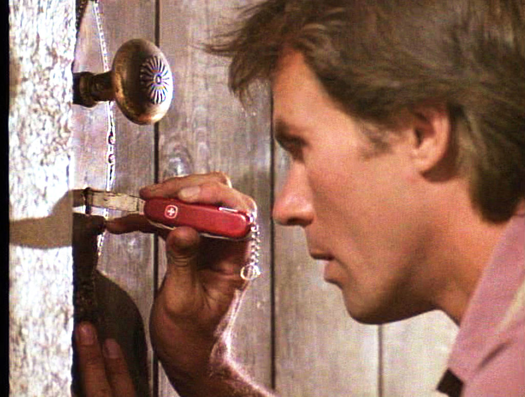 Richard Dean Anderson, wearing casual clothing, uses a Swiss Army knife to work on a door lock in a scene from the 1980s TV series "MacGyver."
