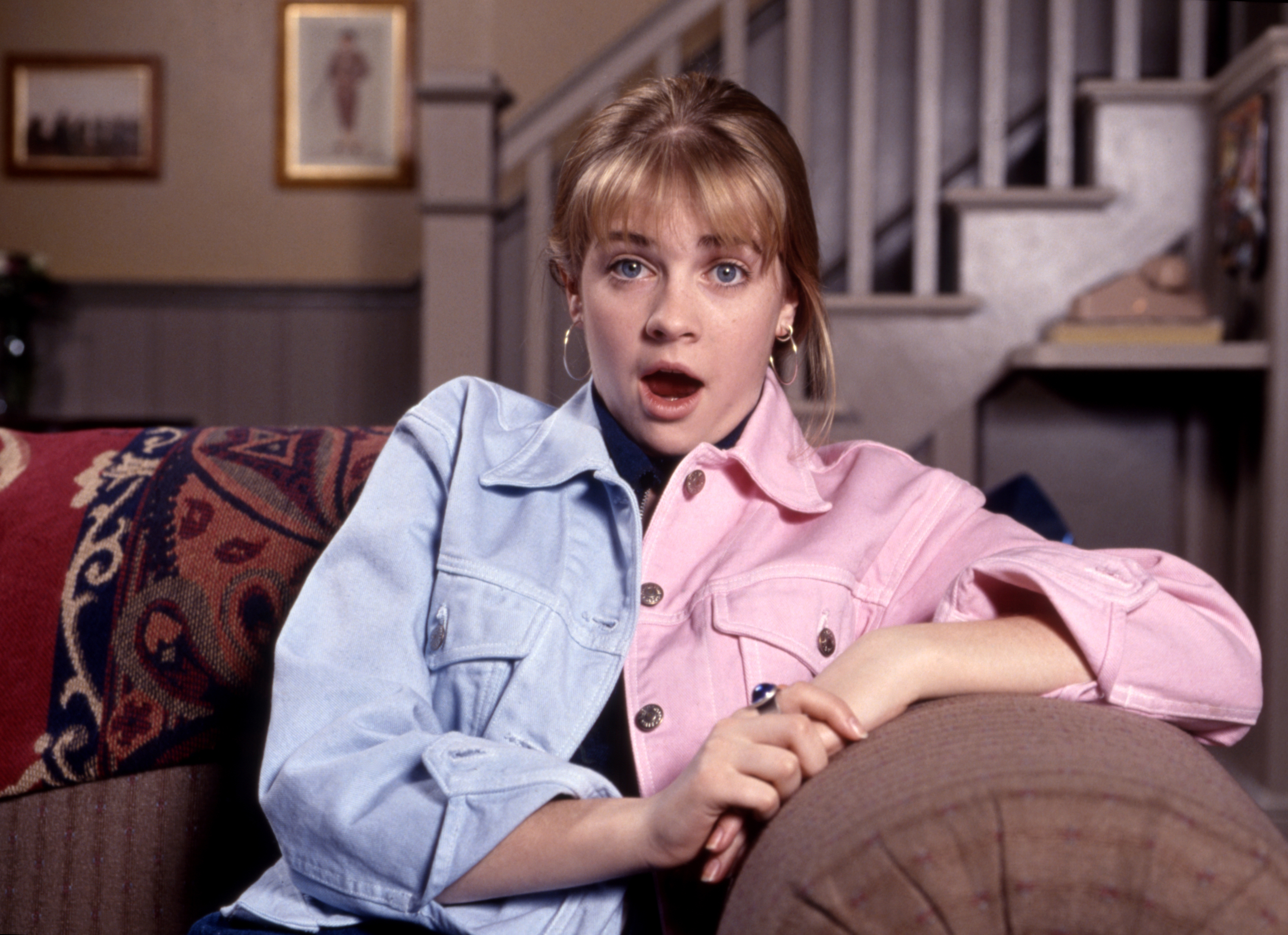 Melissa Joan Hart sits on a couch, wearing a dual-colored denim jacket with a surprised expression