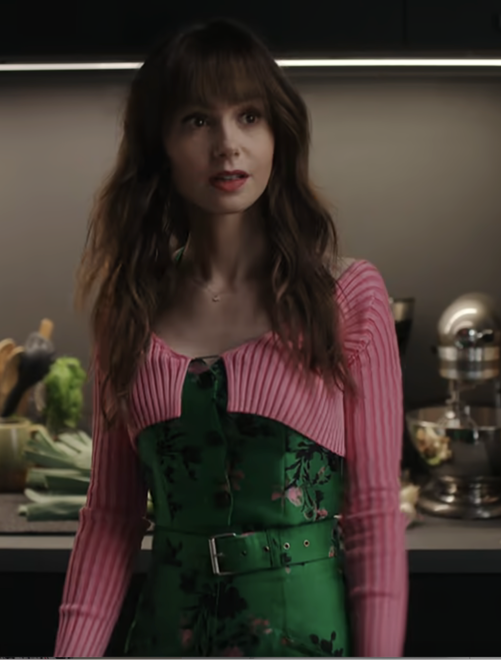 Lily Collins stands in a kitchen, wearing a pink ribbed sweater over a green floral dress with a belt