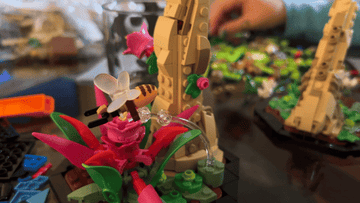 Close-up of a colorful LEGO plant with a LEGO bee and background pieces. A hand is assembling LEGO structures