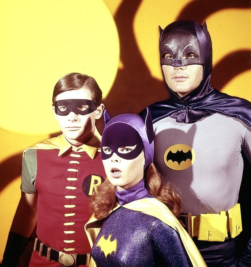 Robin, Batgirl, and Batman in their superhero costumes stand together against a yellow background