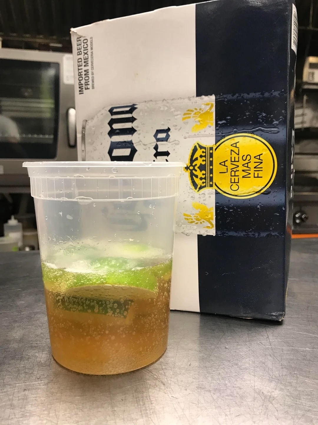 Plastic cup with beverage and lime slices, next to a Corona beer box in a kitchen setting