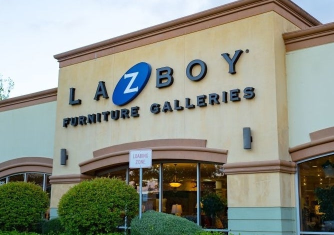 Front view of a La-Z-Boy Furniture Galleries store with its sign prominently displayed. Bushes and trees decorate the exterior, and cars are parked nearby