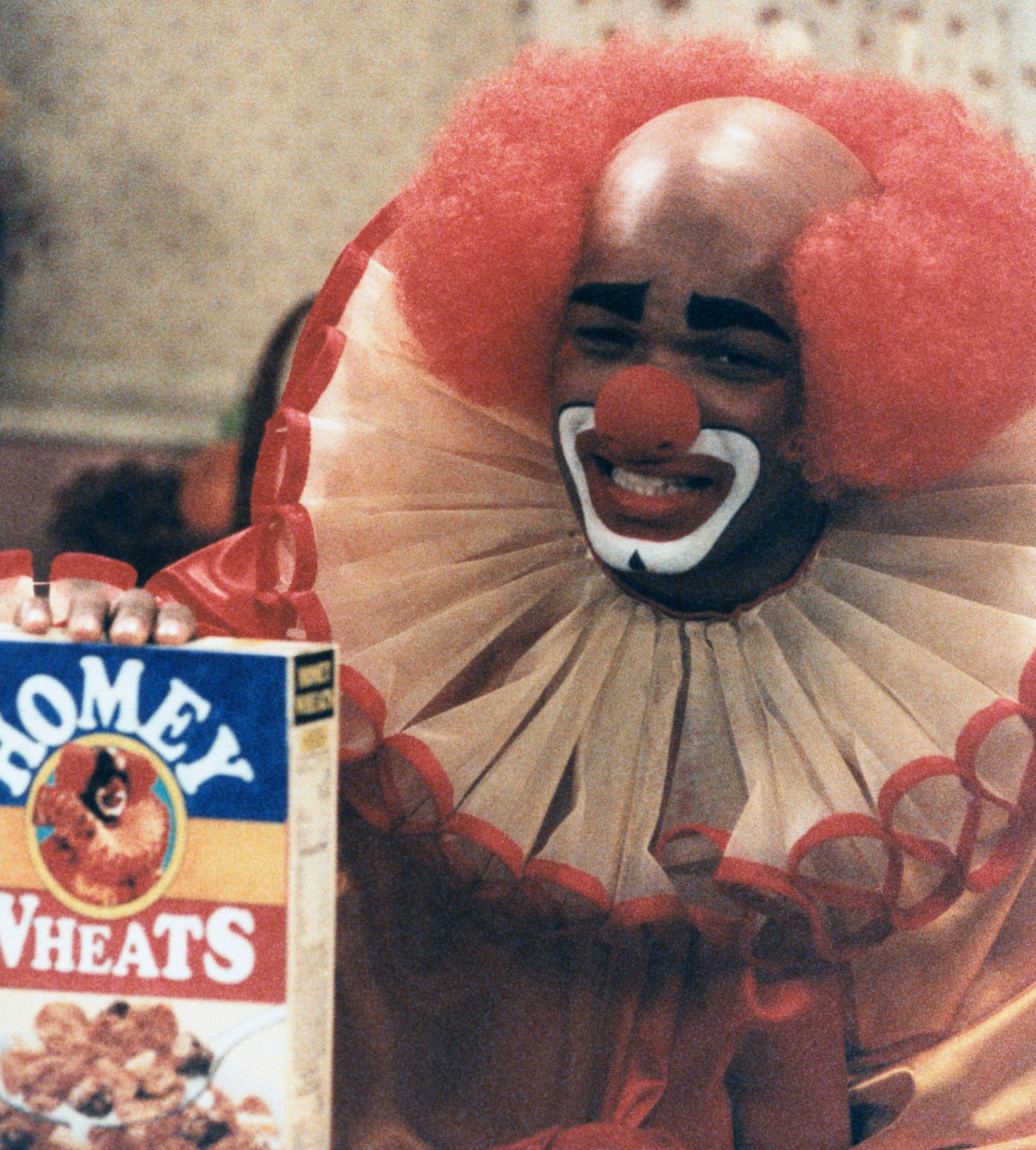 A person dressed as a clown with a painted face, wearing puffy red hair and a ruffled collar, smiles while holding a box of 'Homey Wheats' cereal