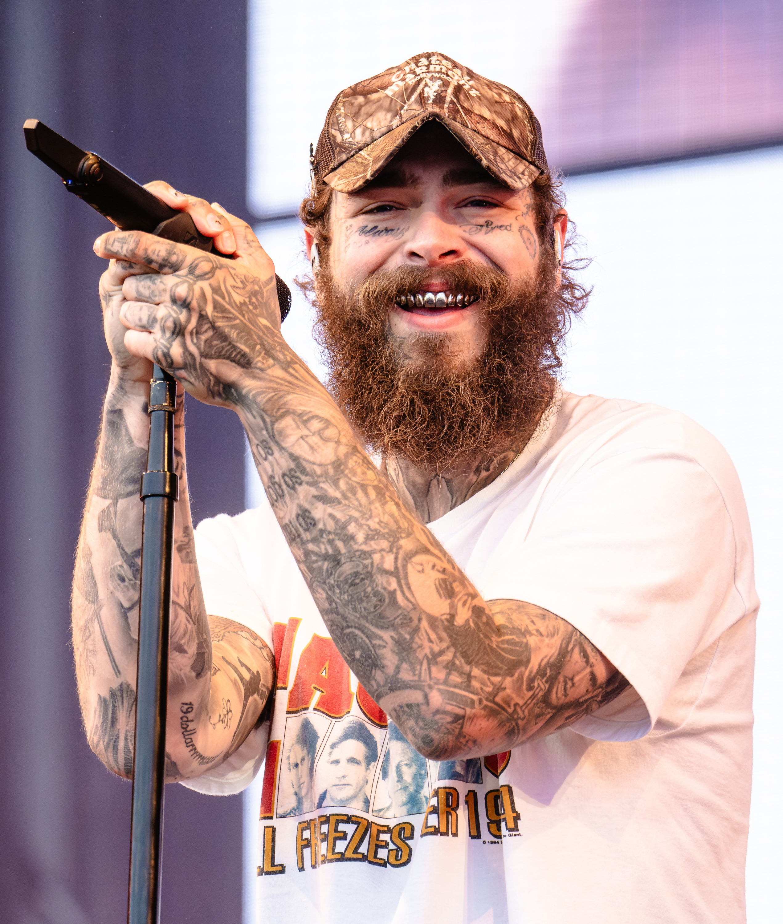 Post Malone performing on stage, wearing a graphic T-shirt, a camouflage baseball cap, and showcasing numerous tattoos on his arms and face