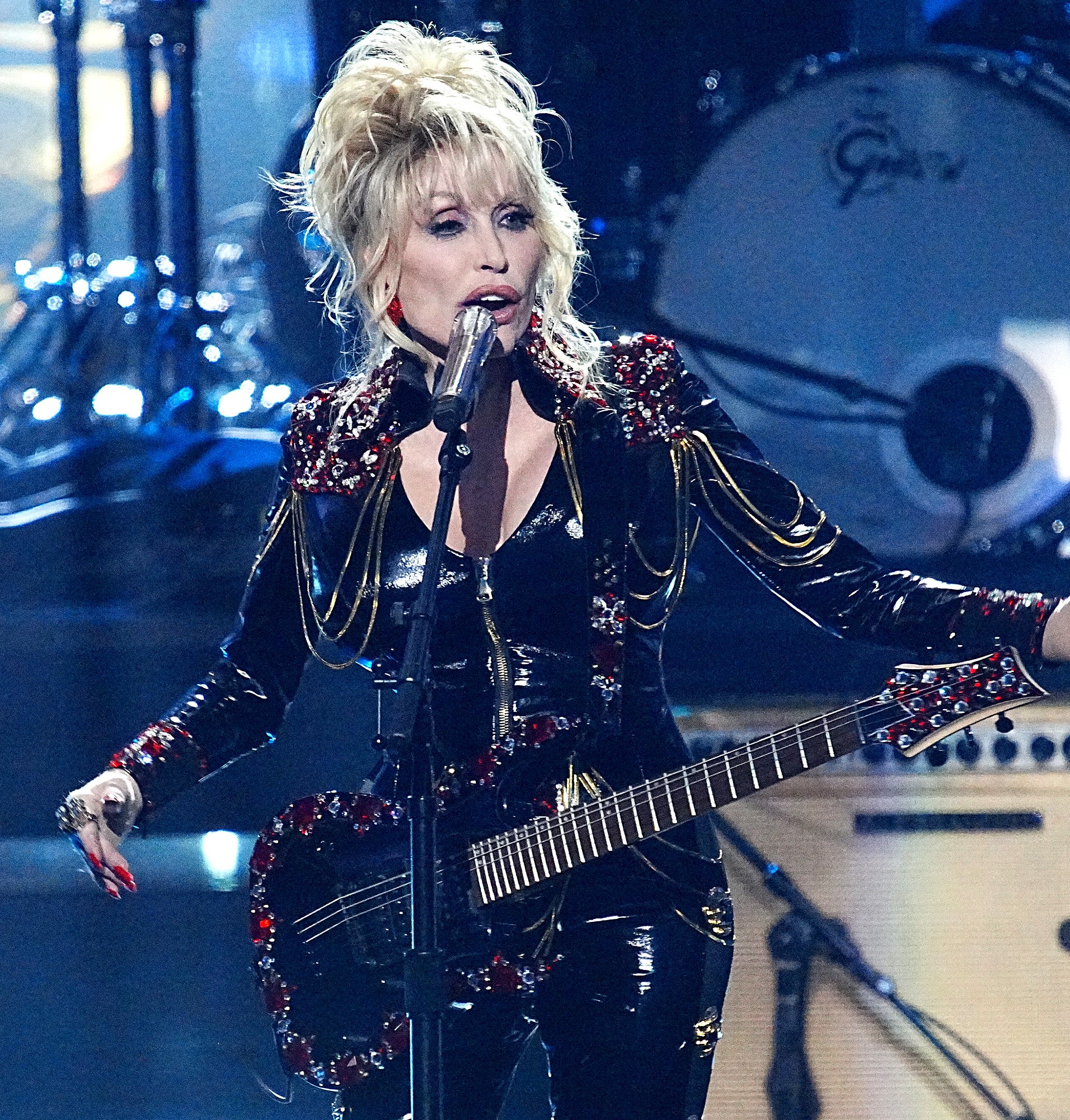 Dolly Parton performs on stage, holding a guitar and wearing a sparkly black outfit with gold and red embellishments, with a drum set in the background