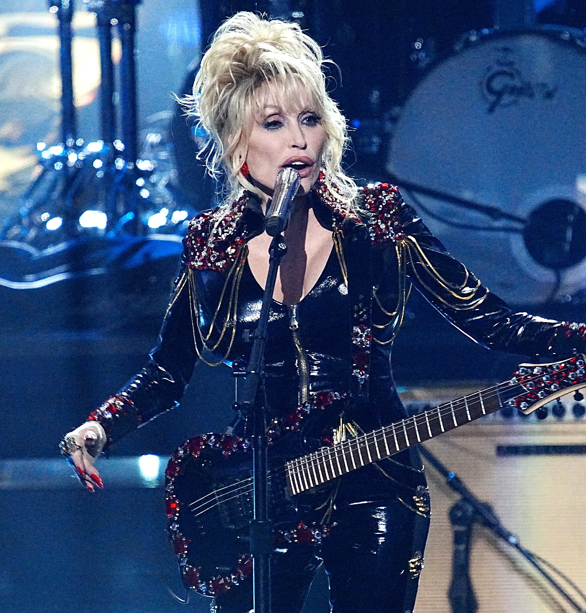Dolly Parton performs on stage, holding a guitar and wearing a sparkly black outfit with gold and red embellishments, with a drum set in the background