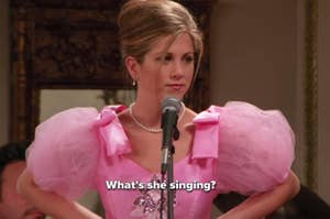Jennifer Aniston dressed in a pink puffy dress with a pearl necklace, stands behind a microphone. Text on the image says, "What's she singing?"