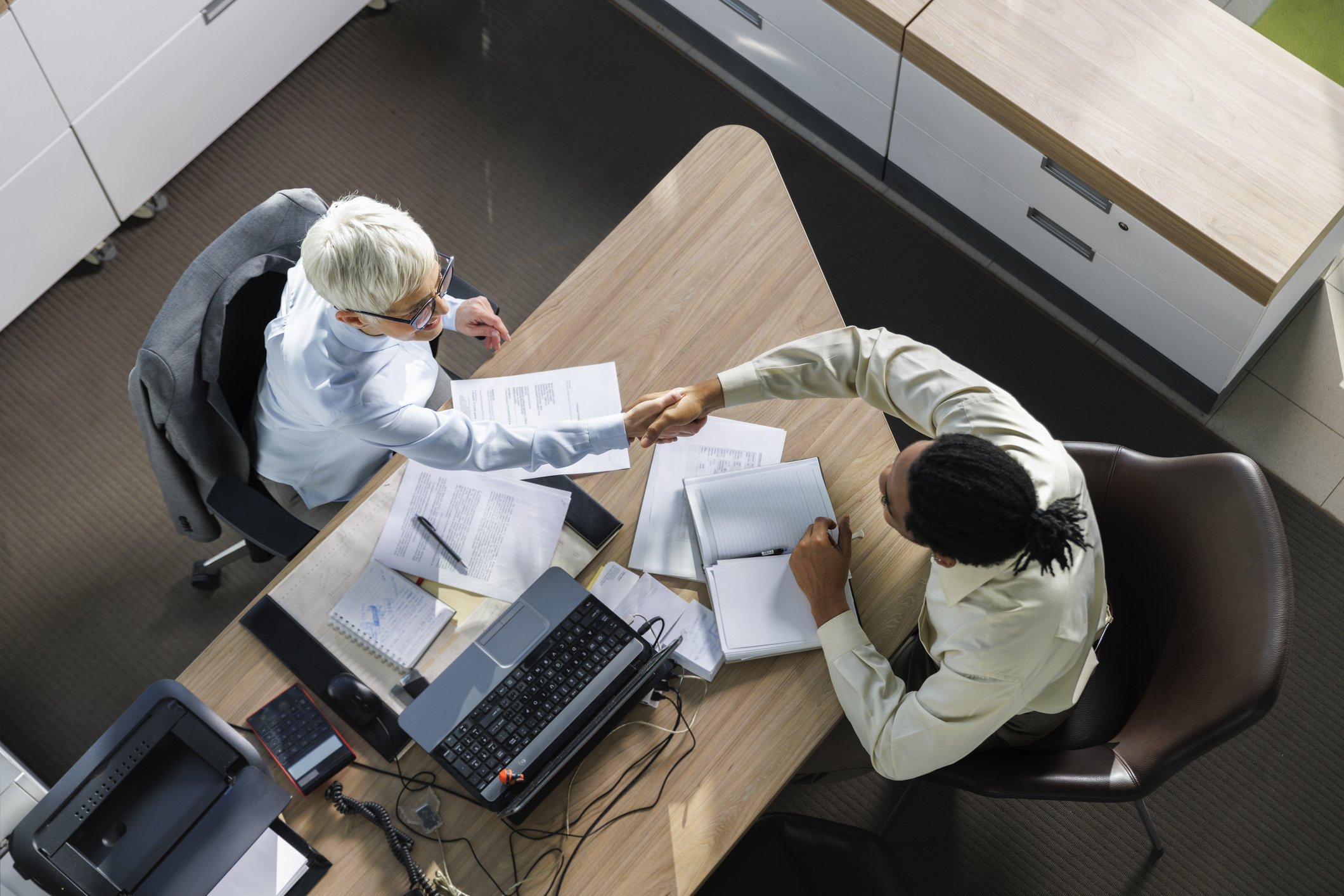 Two individuals, one with short hair and glasses and another with long hair in a ponytail, shake hands across a desk with documents and a laptop