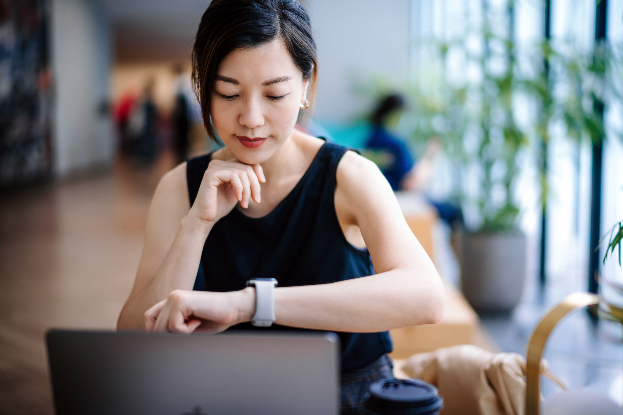 A woman in a sleeveless top works intently on her laptop while checking her smartwatch, with a coffee cup beside her in a modern, brightly-lit workspace