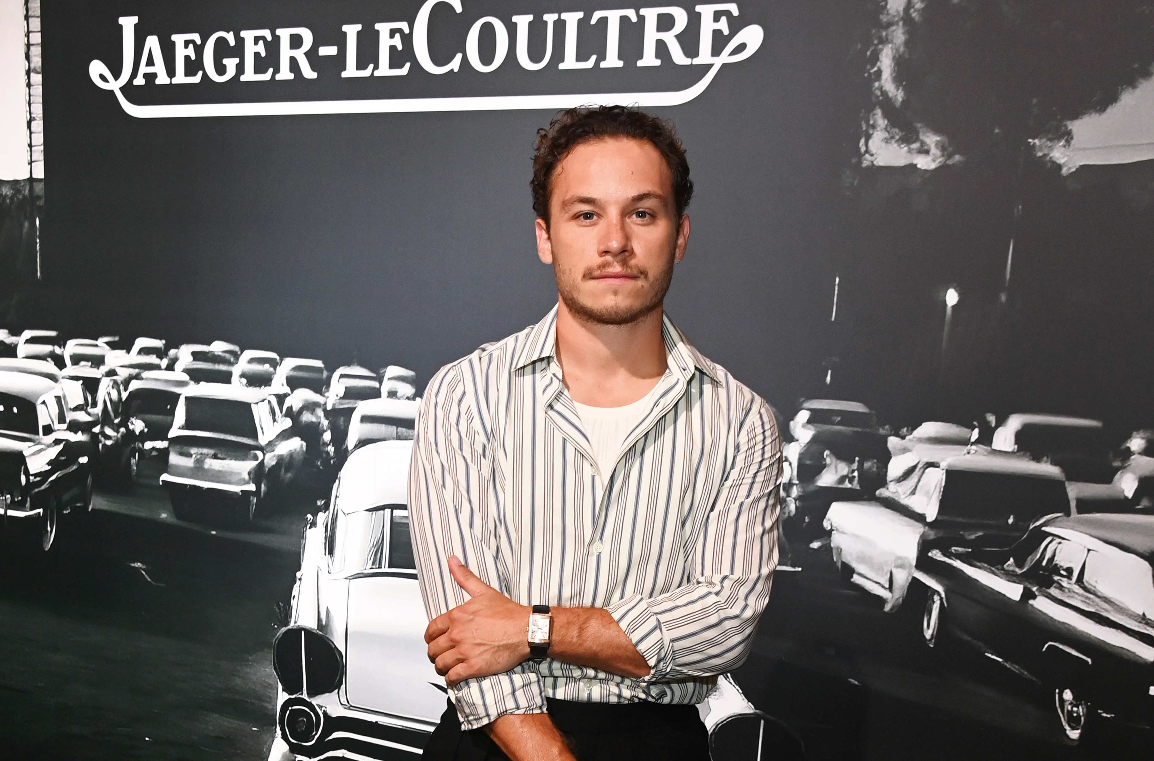 Nick Pugliese poses in front of a vintage car display backdrop at a Jaeger-LeCoultre event, wearing a striped shirt and watch