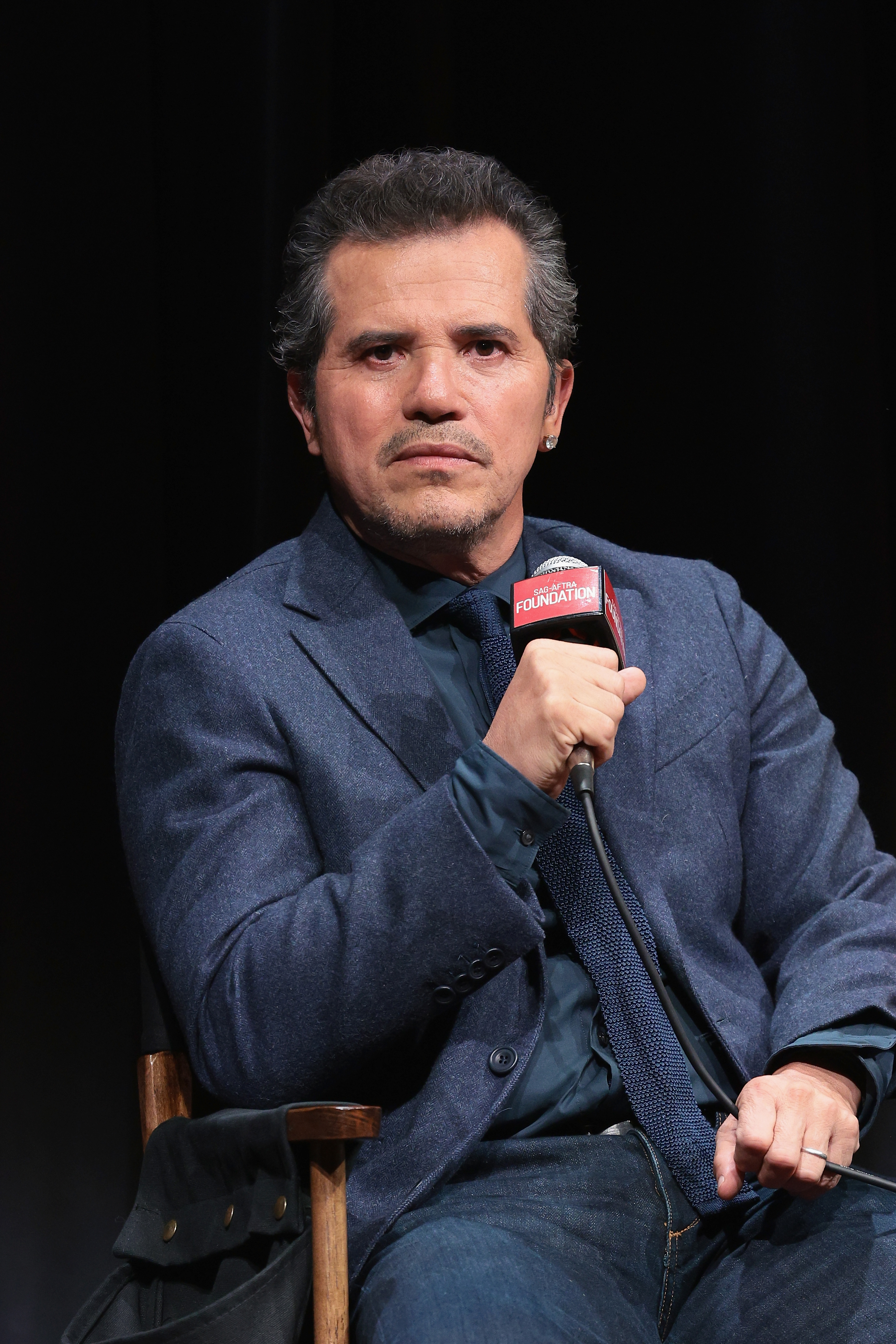 John Leguizamo sits on stage, holding a microphone. He is wearing a blue blazer with a matching shirt and tie