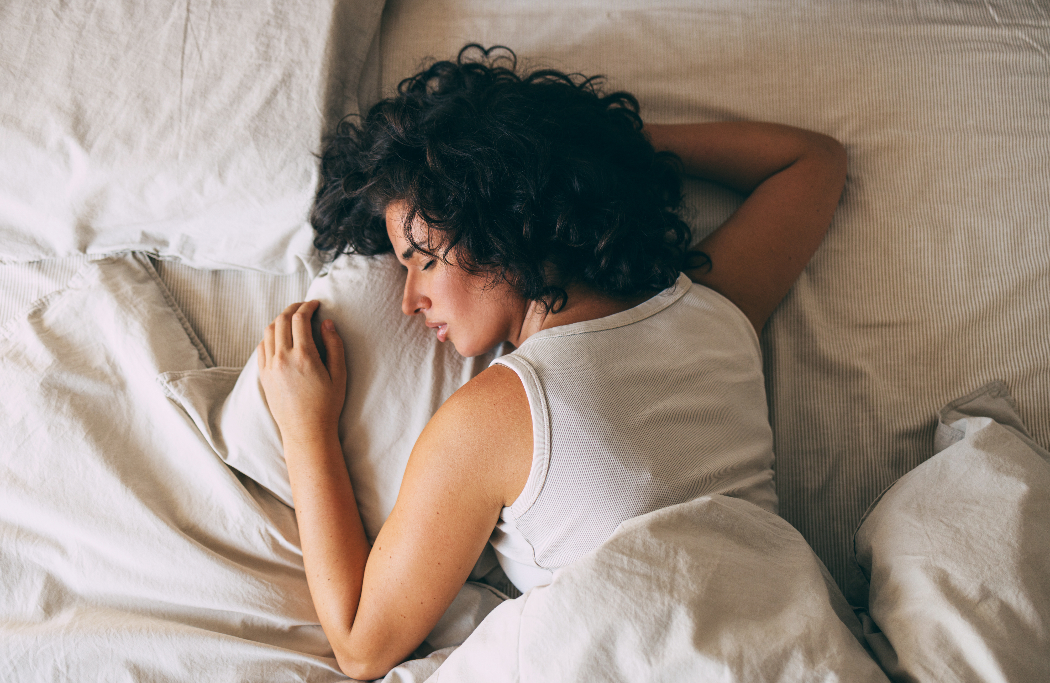 A woman with curly hair is sleeping on her side in bed, wearing a sleeveless top, with her arm resting on the pillow