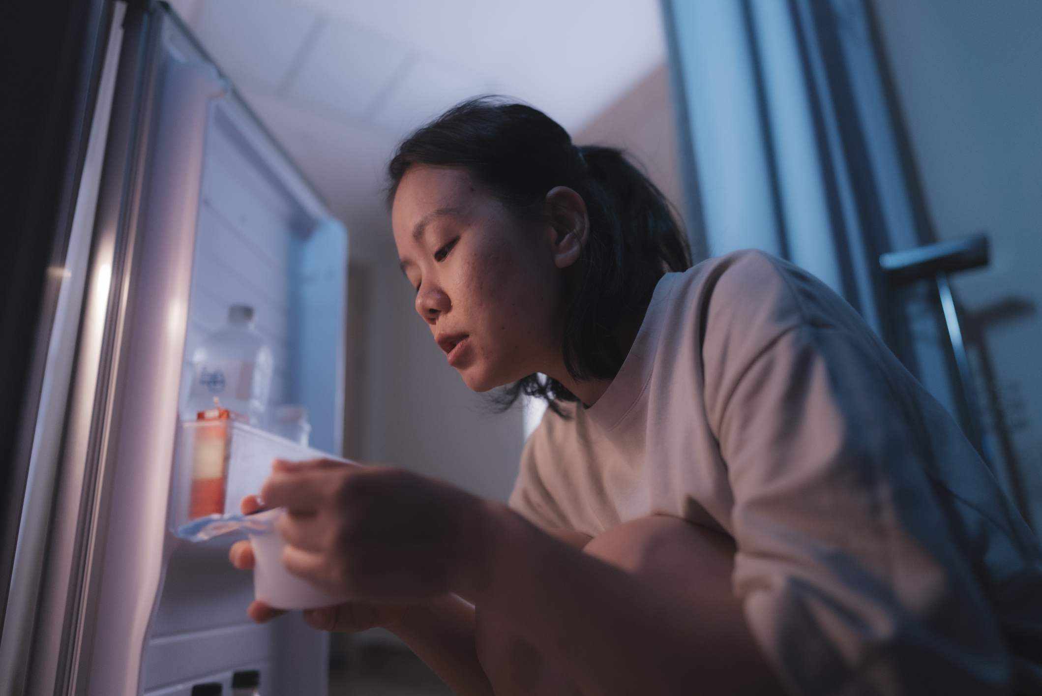A person kneels in front of an open refrigerator, holding a container and looking at it thoughtfully