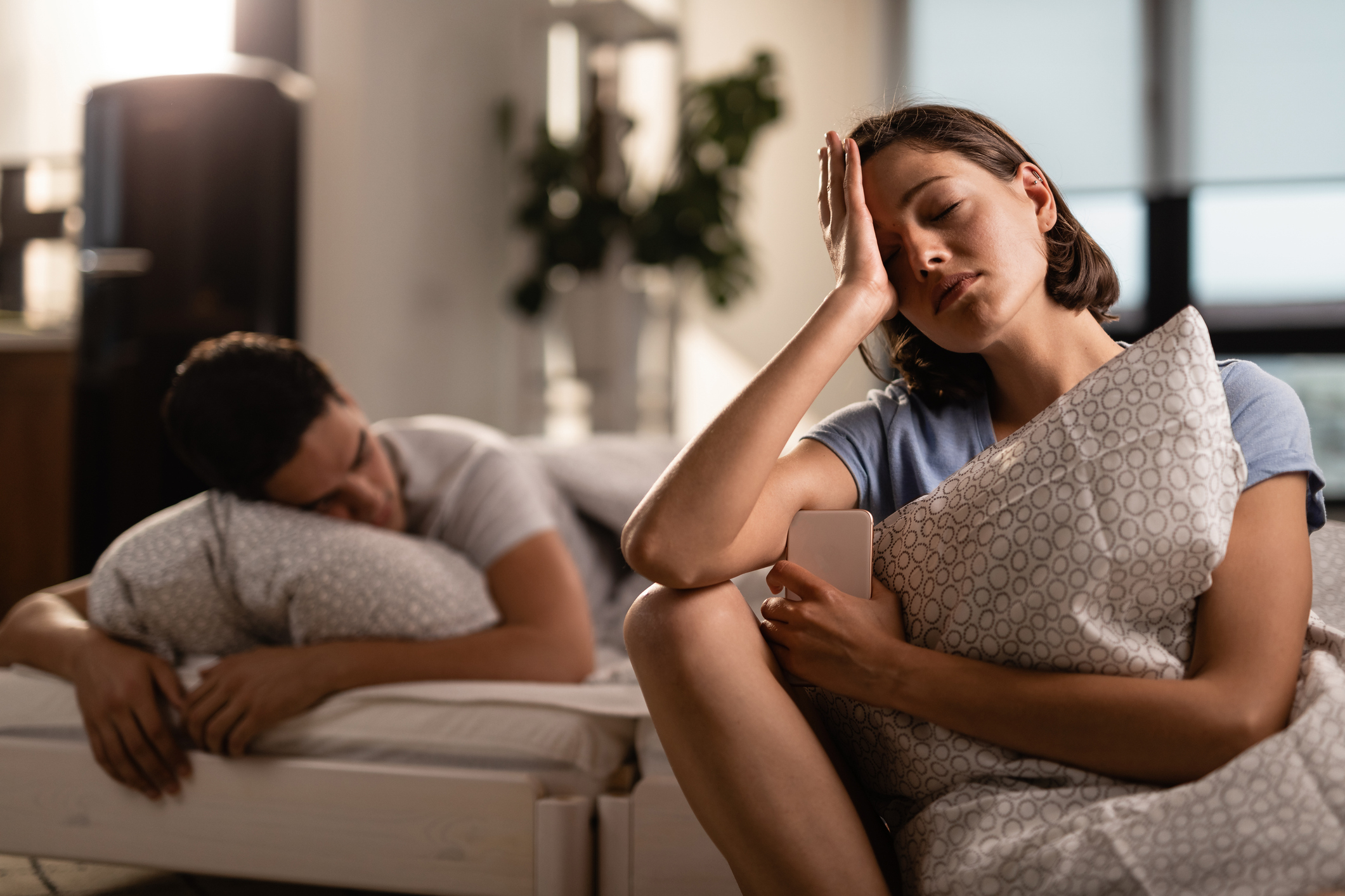 A woman is sitting on a bed, looking stressed while holding a phone, with a man sleeping beside her