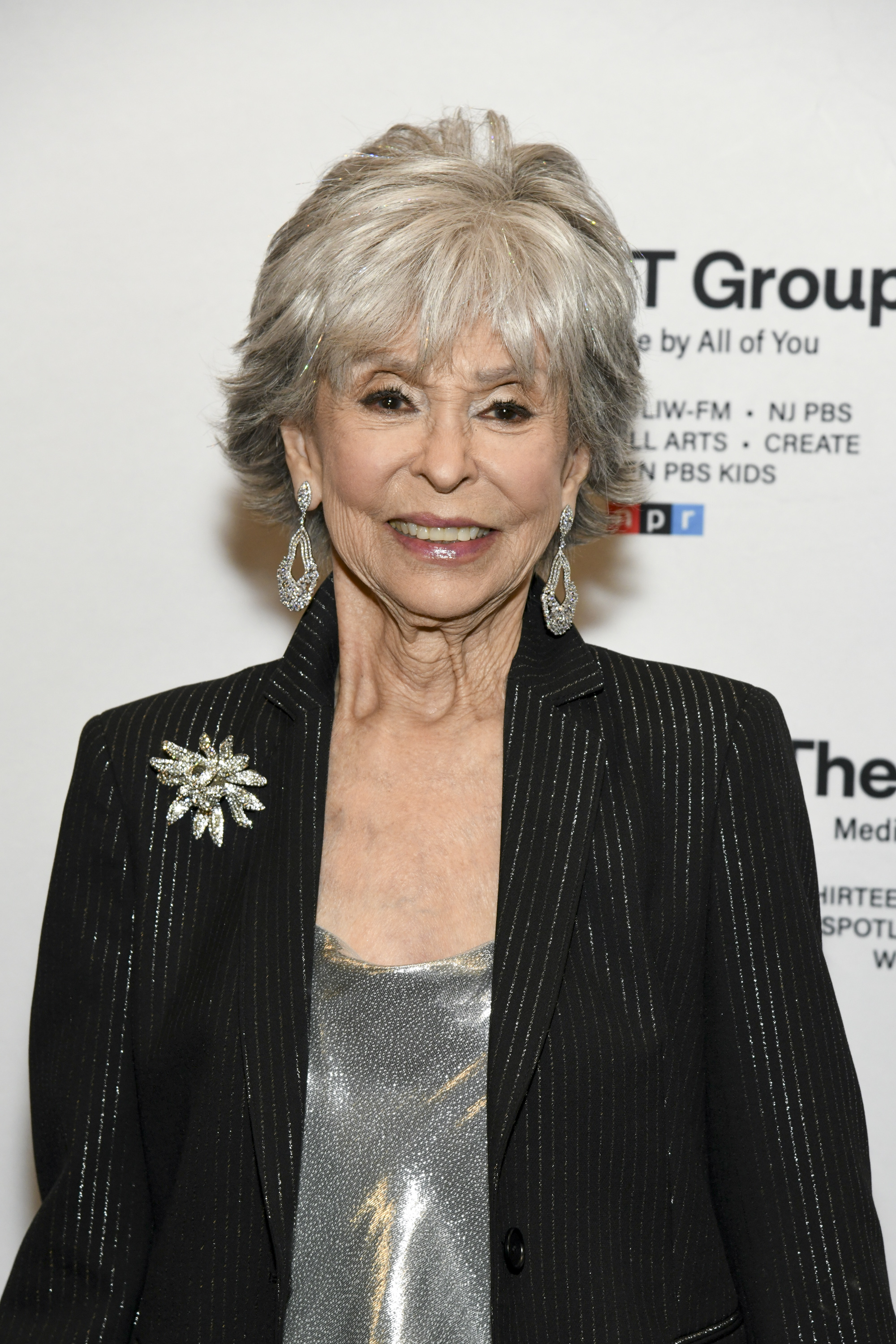 Rita Moreno wearing an elegant pinstriped blazer with a sparkly flower brooch and silver top, attends a formal event