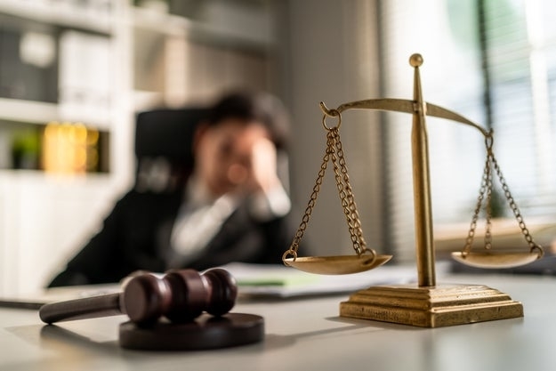 A person is sitting at a desk with their head resting on their hand, looking tired. In the foreground, a gavel and scales of justice are prominently displayed