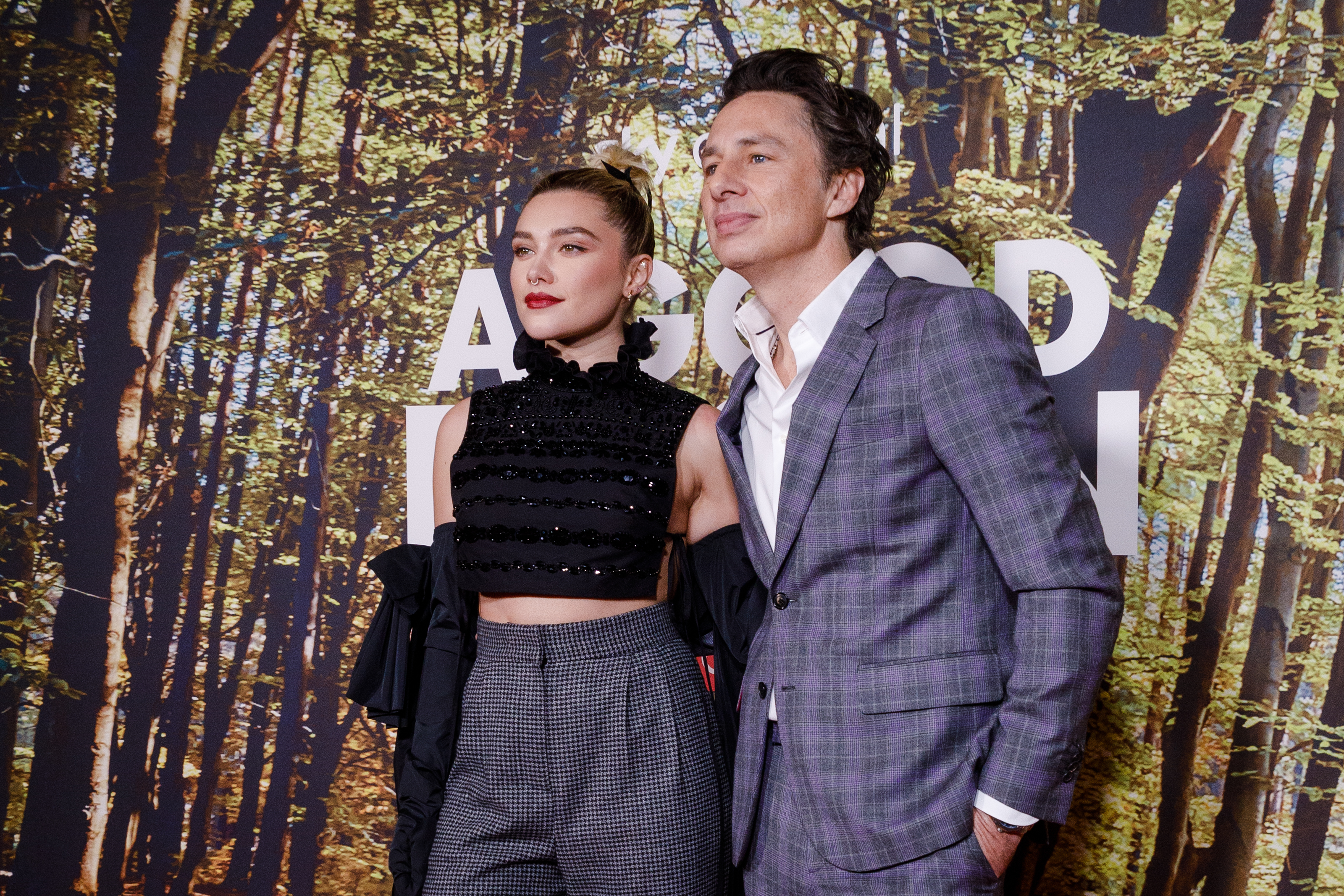 Florence Pugh wearing a stylish top and high-waisted pants, and Zach Braff in a plaid suit at a celebrity event with a forest backdrop
