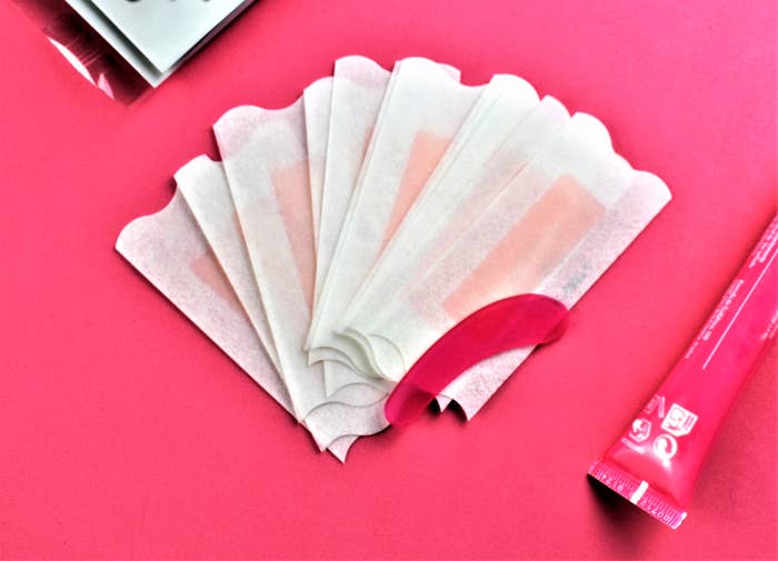 Six wax strips fanned out on a pink surface alongside a pink tube of cream