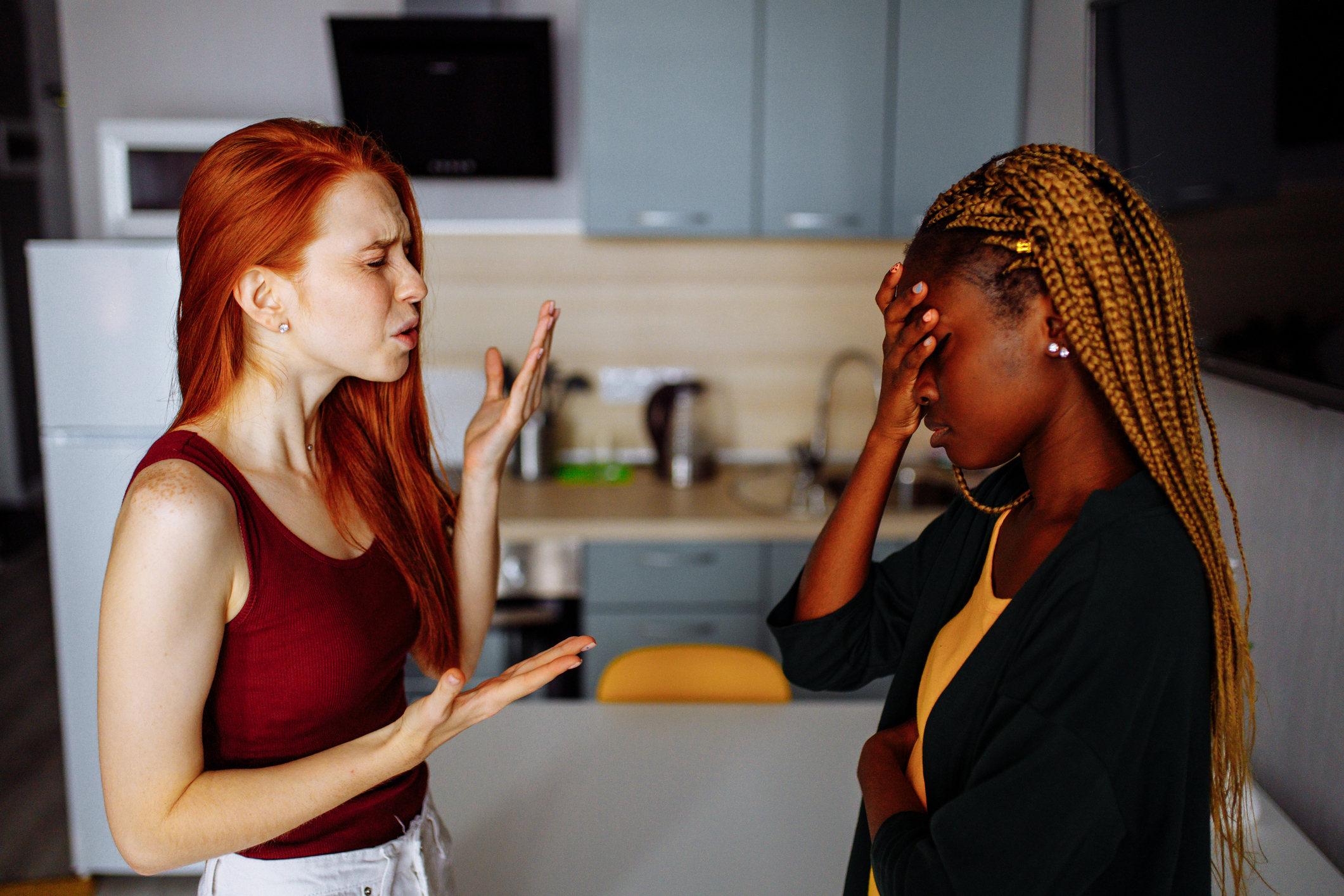 Two women arguing in a kitchen. One has red hair and gestures with frustration while the other, with braided hair, covers her face with her hand