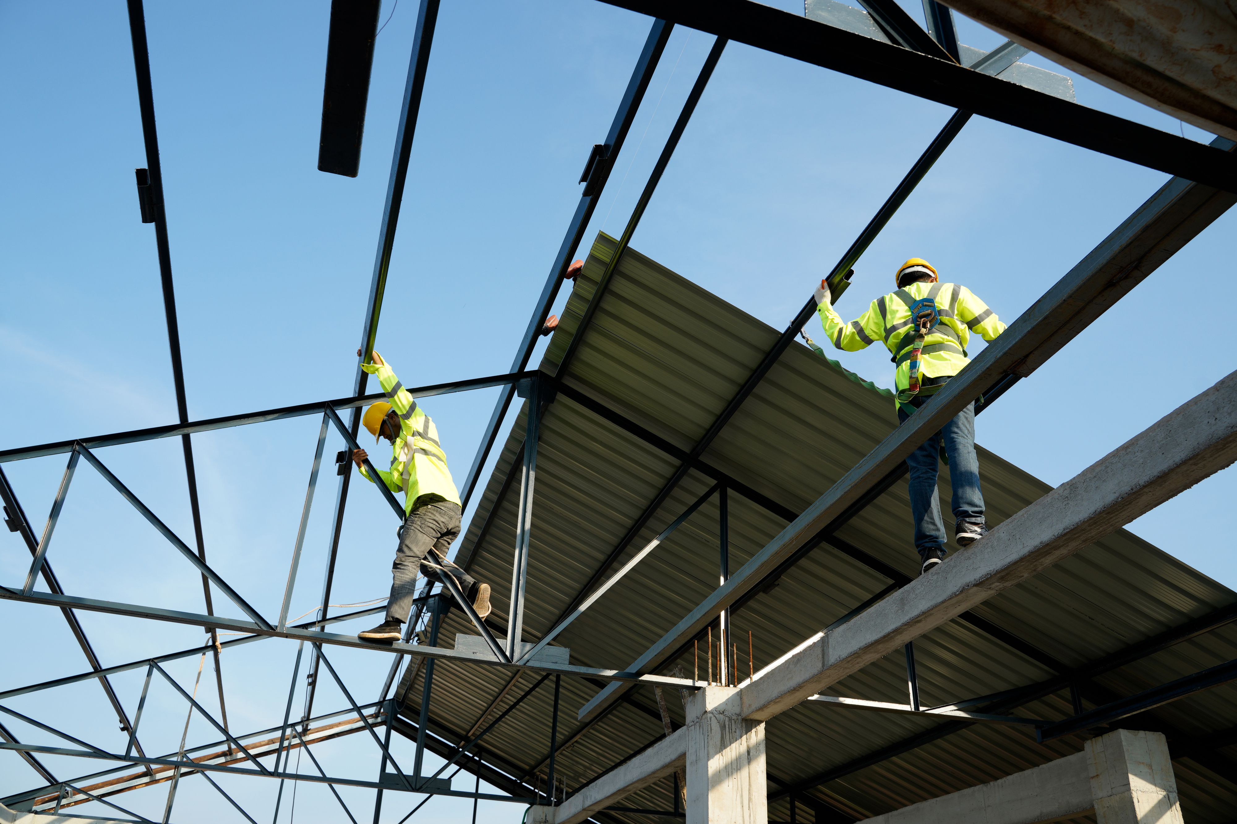 Two construction workers in safety vests are installing roof beams on a building framework at a construction site