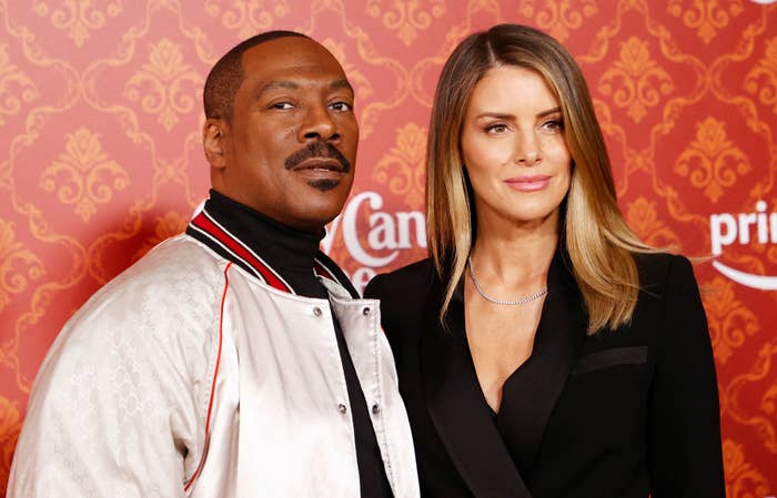 Eddie Murphy in a casual jacket and Paige Butcher in a stylish black outfit pose together on a red carpet event backdrop