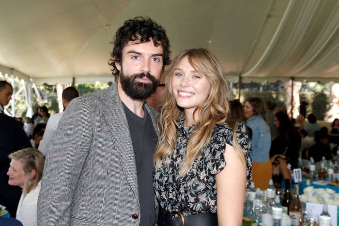 Robbie Arnett and Elizabeth Olsen pose together under a tent at an event. Robbie wears a casual blazer; Elizabeth is in a floral-print blouse