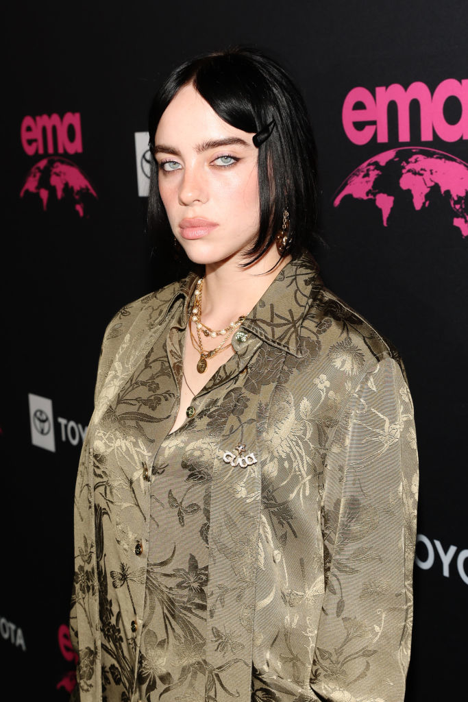 Billie Eilish stands on the red carpet, wearing a patterned shirt and layered necklaces, at the EMA event
