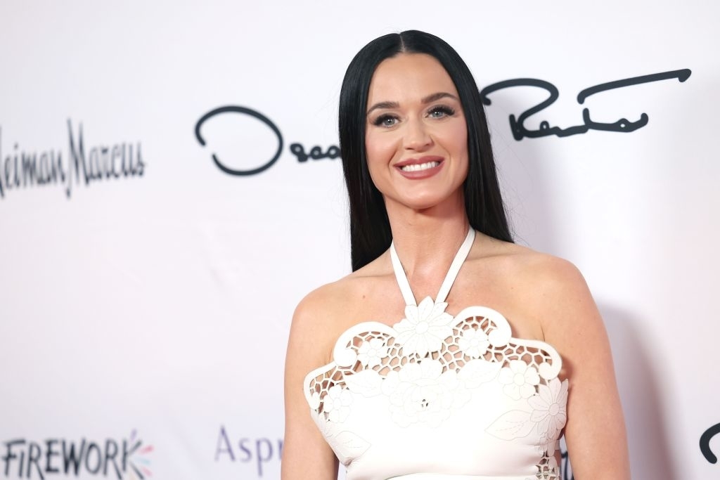 Katy Perry on a red carpet, wearing a sleeveless white gown with intricate cut-out details at the neckline